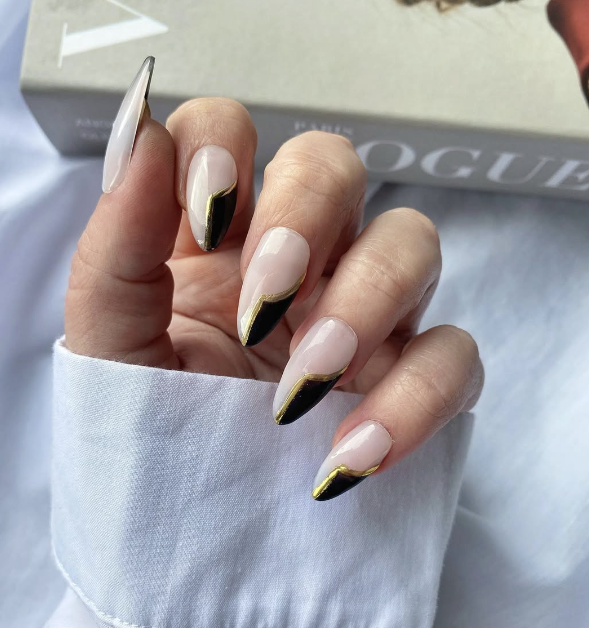Long almond-shaped nails with a nude base, a sharp asymmetric black French tip, and a metallic gold line tracing the inner edge.