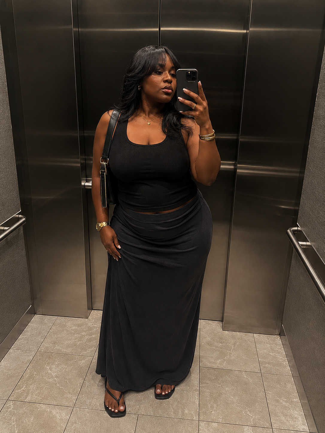 A person taking a mirror selfie in an elevator wearing a fitted black tank top, a low-waisted dark grey maxi skirt, and flat black flip flops.