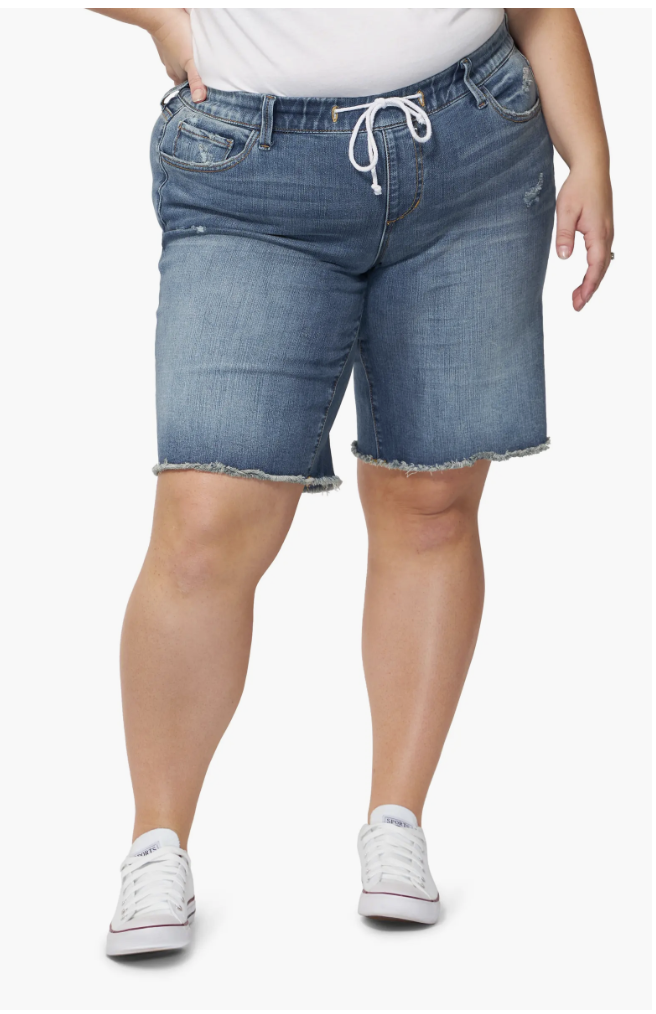 A woman wearing medium-wash SLINK Jeans frayed hem denim Bermuda shorts, featuring a curvy fit designed to stay in place on thick thighs and support an apron belly.