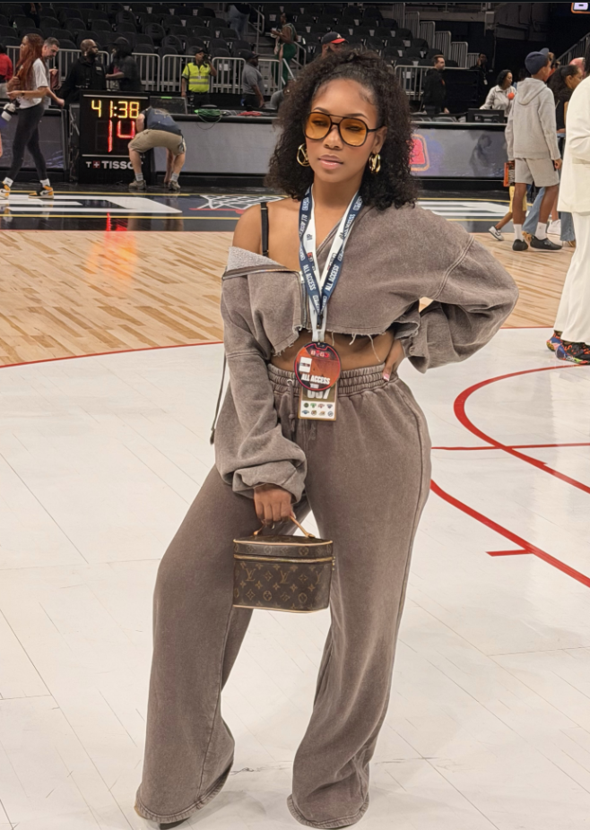 Woman courtside wearing a taupe off-the-shoulder sweatsuit with a small structured brown handbag.