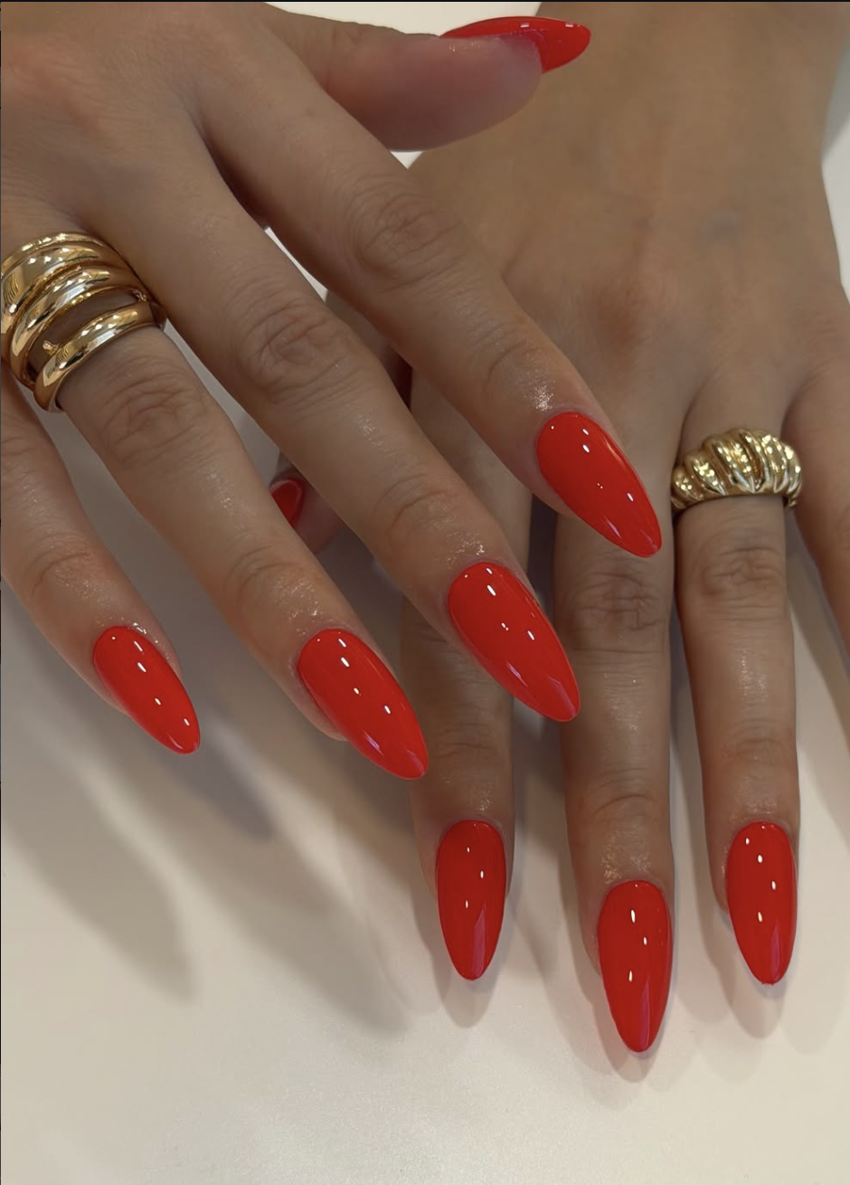 A close-up of long, stiletto-shaped nails painted in a glossy, high-saturation classic bright red, styled with multiple thick, stacked gold rings.