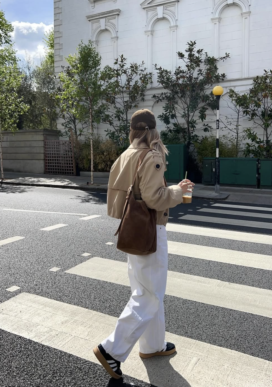 A woman walking across a street in white wide-leg jeans paired with black and white Adidas Breaknet Sleek sneakers and a tan jacket.