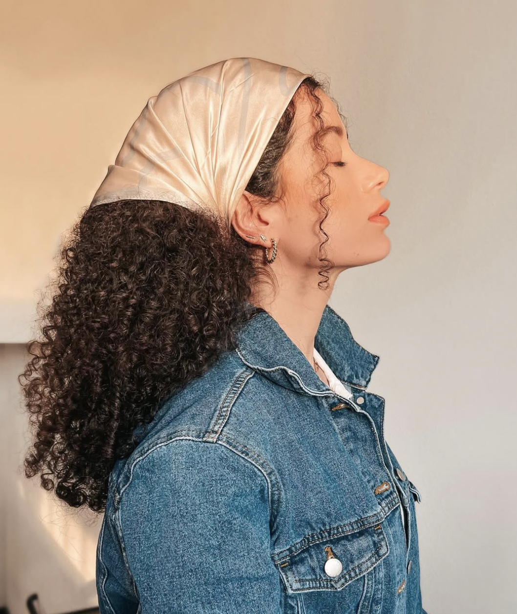A woman in profile wearing a cream-colored silk headscarf tied as a wide headband wrap over long, dark curly hair, paired with a denim jacket.