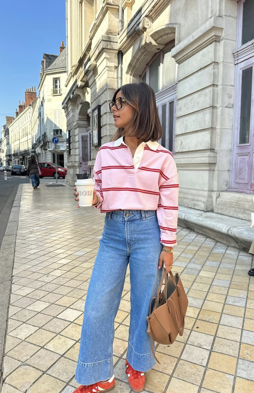A woman wearing a pink and white striped long-sleeve polo sweater tucked into high-waisted blue jeans, holding a coffee cup while walking on a city sidewalk.
