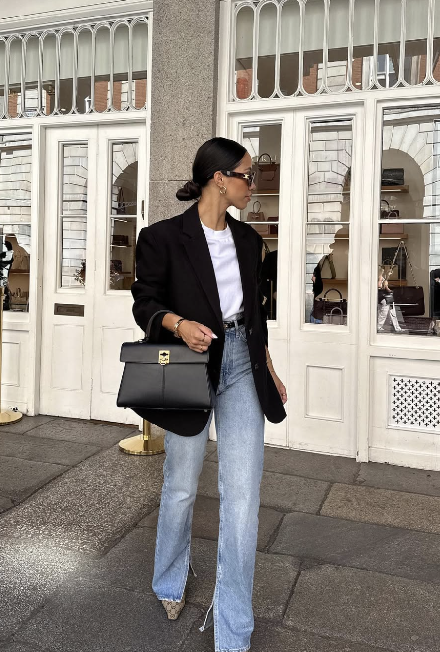 A woman wearing light-wash straight-leg jeans, a white t-shirt, a tailored black blazer, sunglasses, and holding a black top-handle handbag outdoors.
