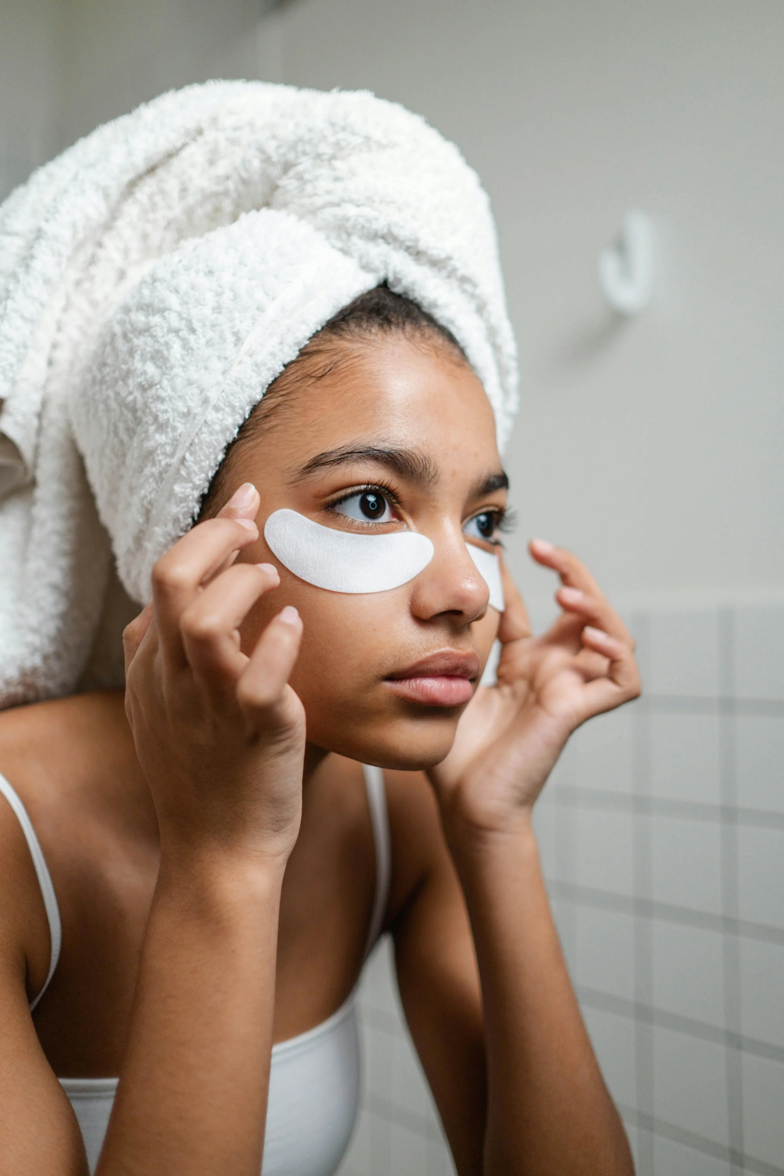 A close-up of a woman wearing gold under-eye patches while looking into a mirror, representing a morning skincare routine for de-puffing and brightening.
