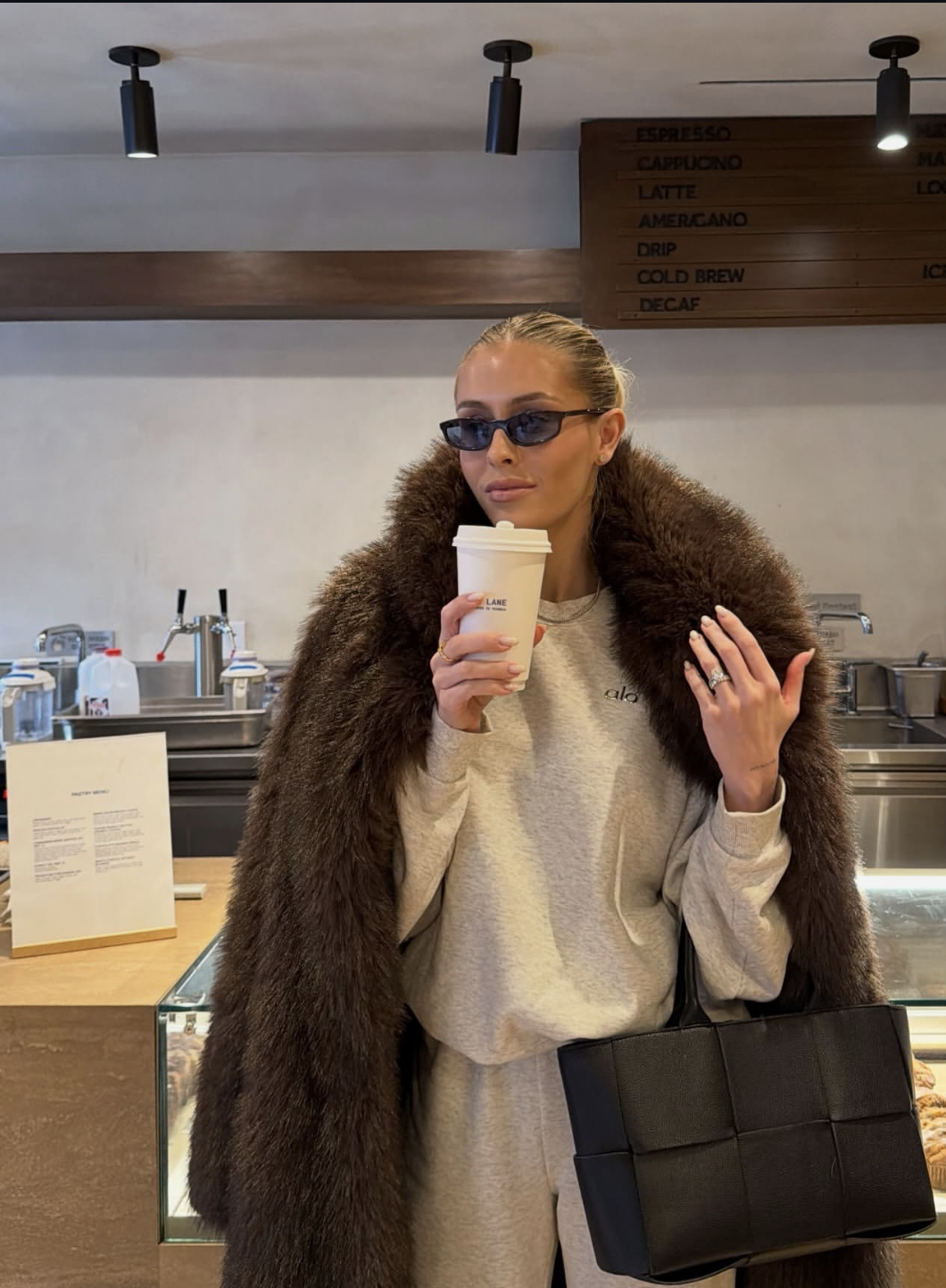 Brown furry-textured coat over a pale sweatshirt and matching-looking trousers with a large dark woven-look tote in a café.