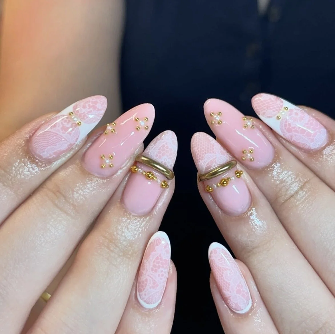 Long almond-shaped pink nails featuring a delicate white lace pattern, small gold stud accents, and chunky gold rings.