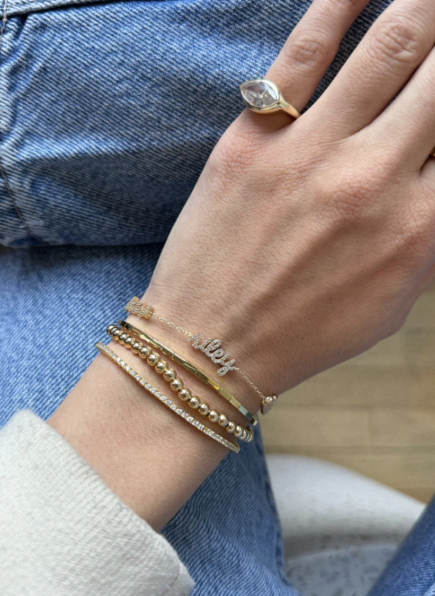 A person wearing a stack of gold jewelry including a Baublebar 14K gold and pavé custom script multi-name bracelet.