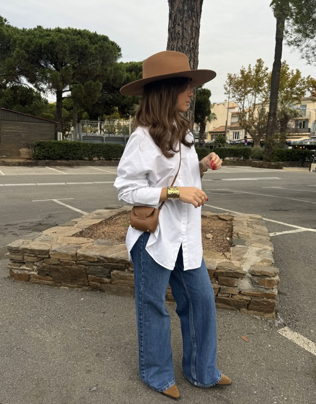 A woman wearing relaxed blue jeans, an oversized white button-down shirt, brown shoes, and a small brown crossbody leather bag.
