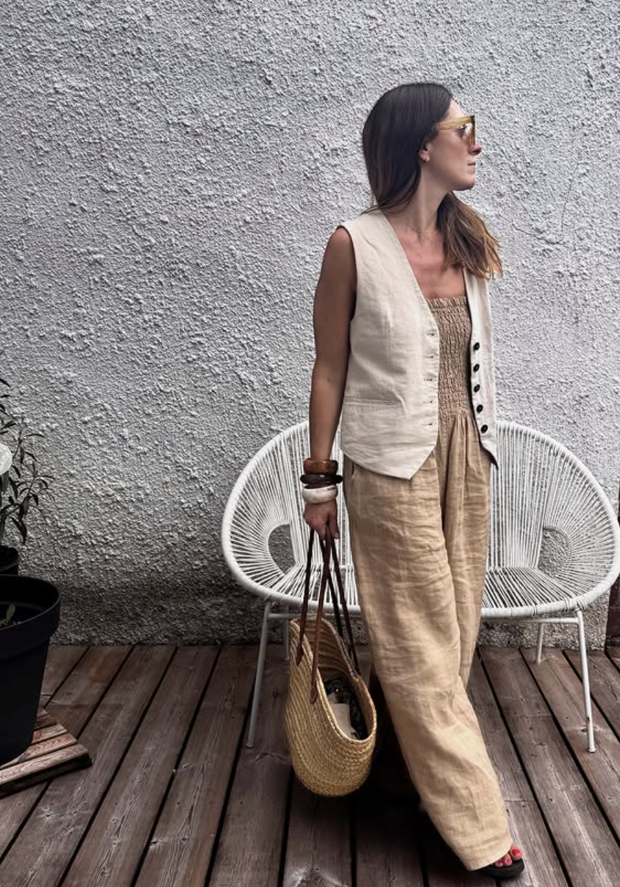 A person wearing a neutral linen tailored vest and matching wide-leg trousers, carrying a large woven straw shoulder bag next to a white round chair.