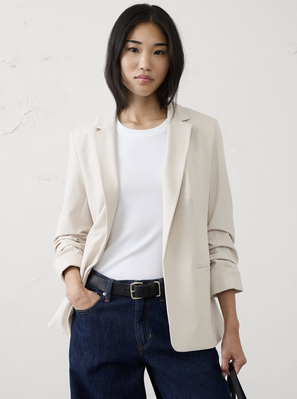 A woman wearing a beige soft-shoulder drape blazer over a white shirt and dark denim jeans with a black belt for a chic office look.