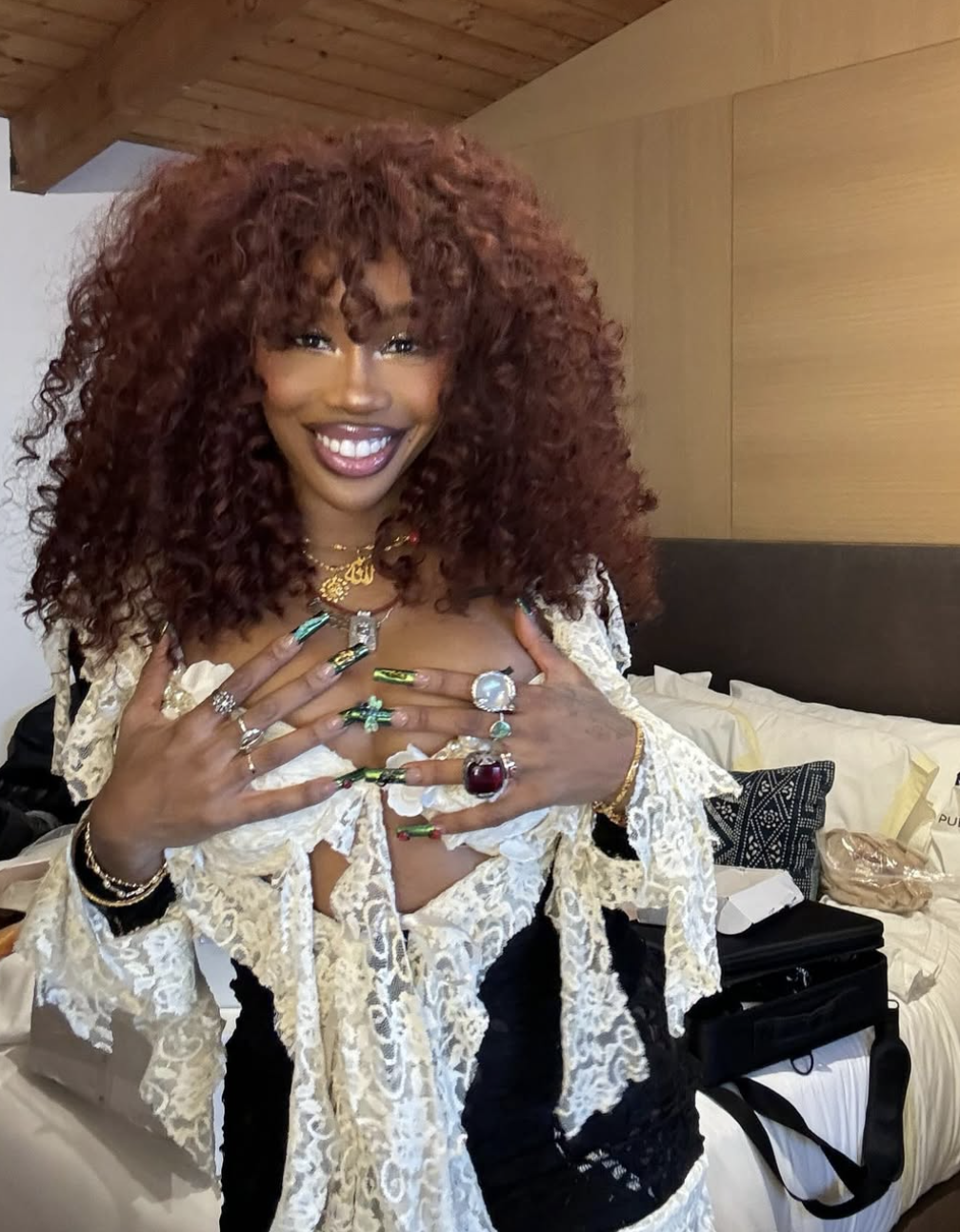 SZA wearing a voluminous dark auburn wavy hairstyle with honey-toned highlights, styled with an intricate lace top and statement rings.