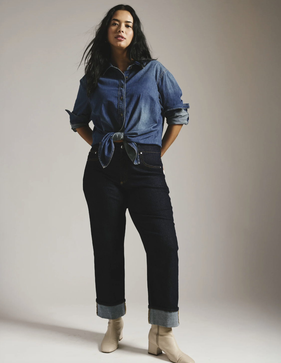 A person with an hourglass figure wearing dark wash Universal Standard Stevie high-rise straight-leg jeans with a thick cuff, styled with a denim button-down shirt tied at the waist to highlight the silhouette.