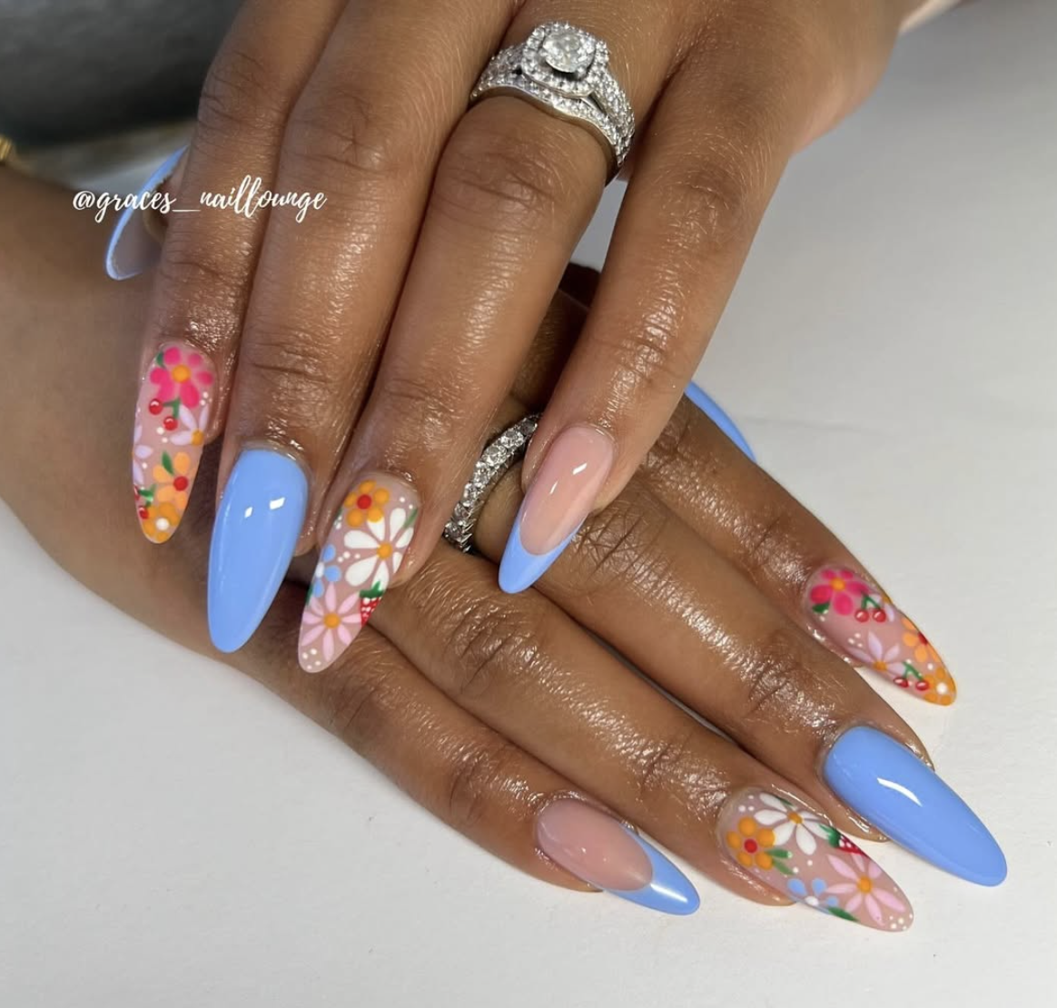High-feminine almond nails by @graces_naillounge featuring periwinkle blue French tips and clear accent nails with bright pink, orange, and white hand-painted flowers.