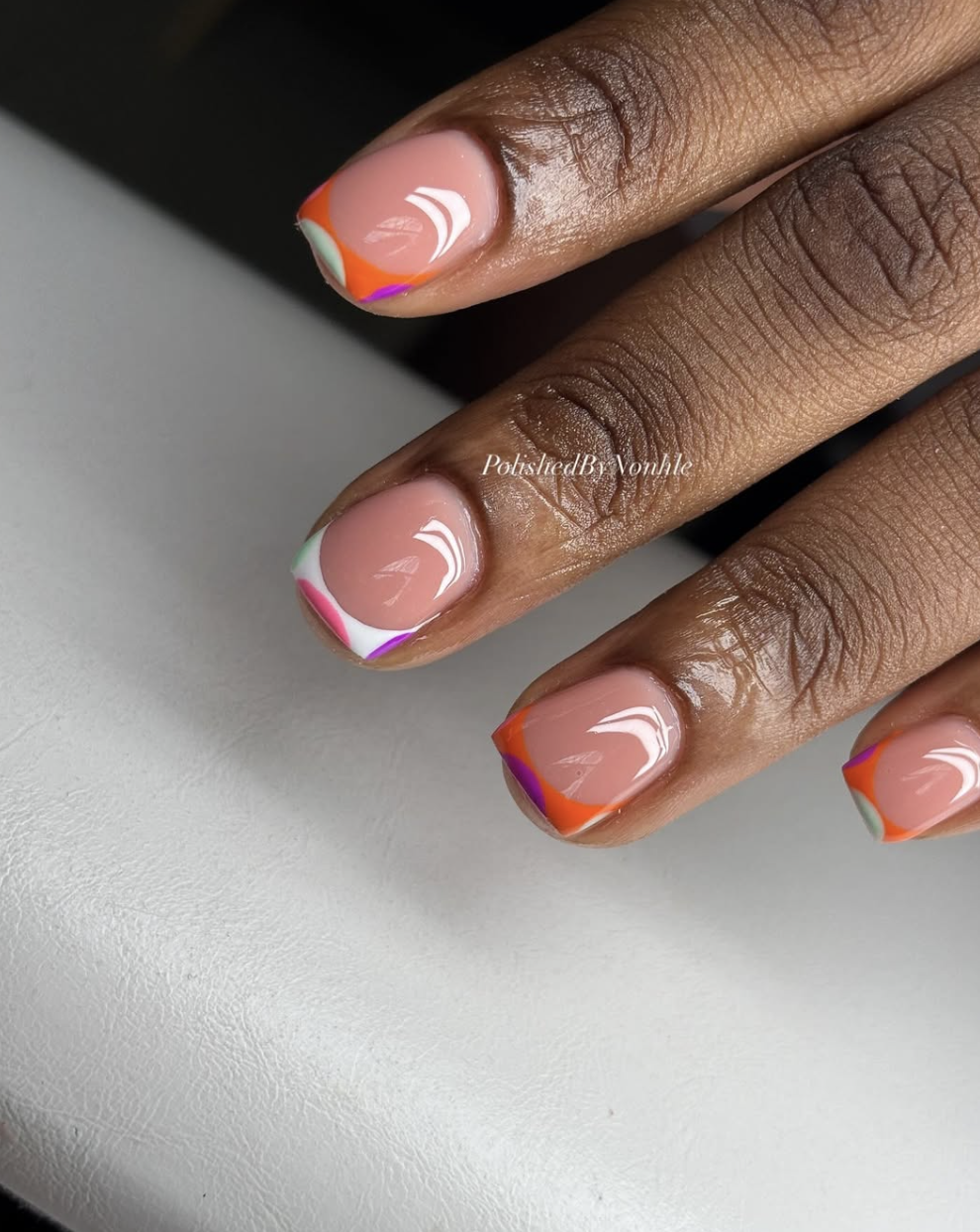A close-up of short nails featuring an asymmetrical, multi-colored diagonal French tip design with pink, orange, and purple swirls over a nude base.