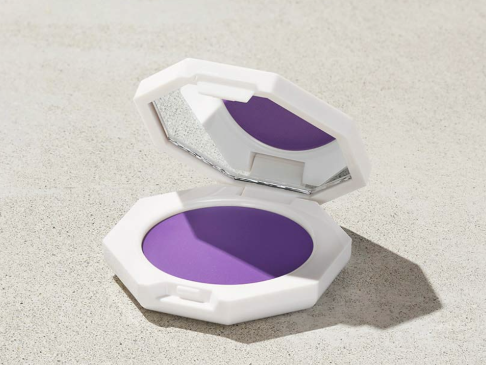 An open white hexagonal compact of Fenty Beauty Cheeks Out Freestyle Cream Blush in the shade Drama Cla$$, a bright violet-purple cream blush with a smooth finish.