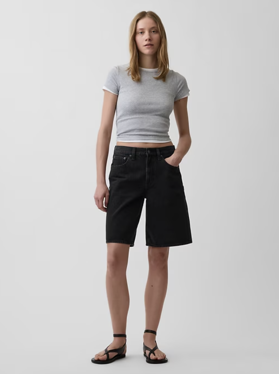 A woman wearing black Gap 11-inch mid-rise longline denim Bermuda shorts with a raw hem, styled with a grey t-shirt and black sandals.
