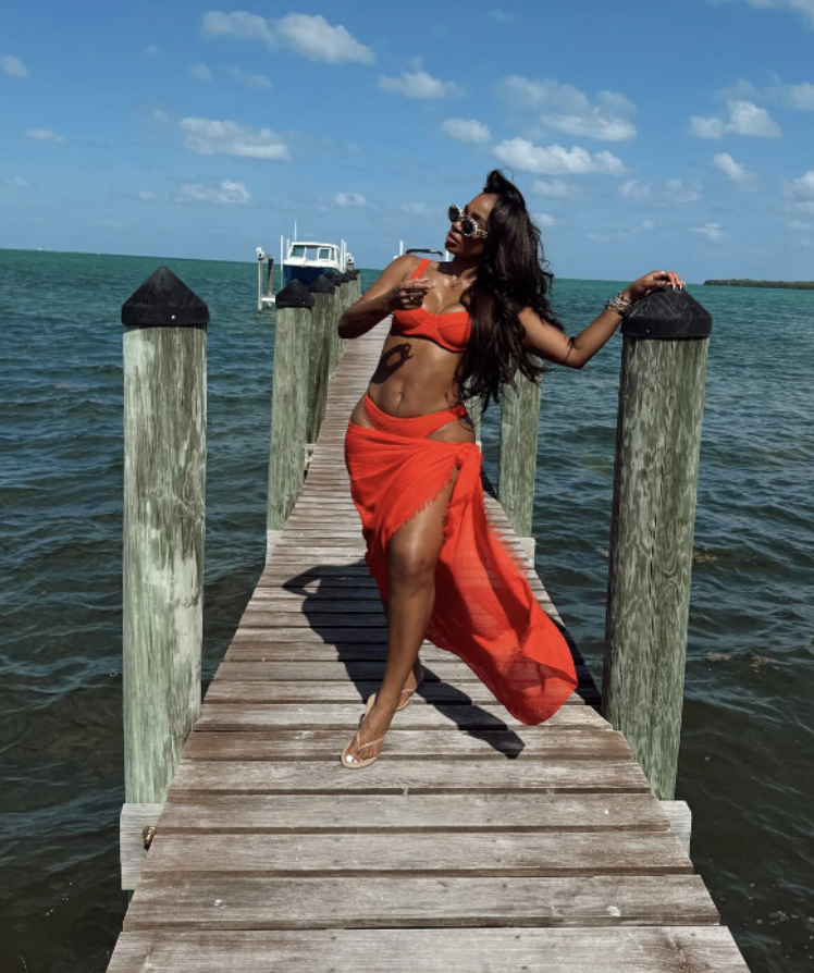 A woman wearing a black tank dress and white slides standing on a wooden pier by the water, illustrating a casual summer BBQ outfit.