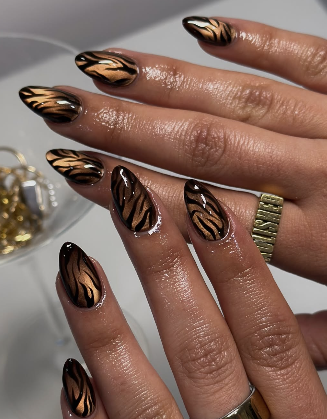 A close-up of almond-shaped nails featuring a magnetic brown cat-eye base with shimmering metallic copper tiger stripes and swirl nail art.