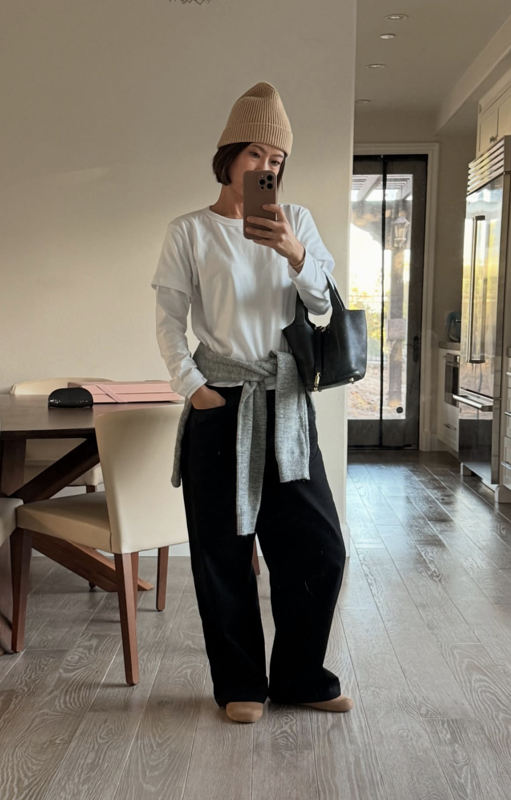 A woman wearing a white layered henley long-sleeve top with a tan beanie, black pants, and a grey sweater tied around her waist.