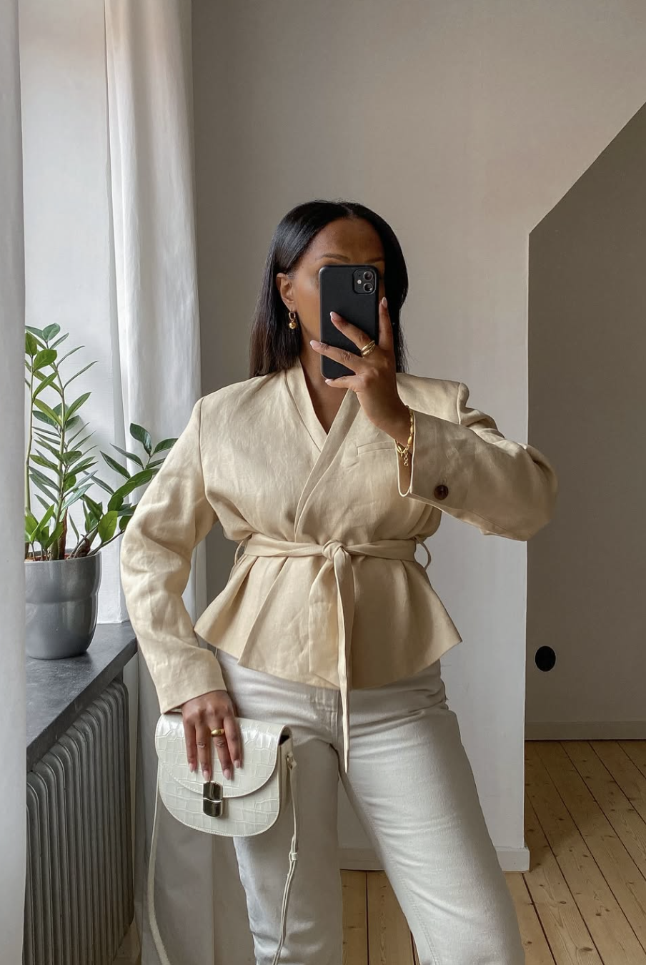 A woman taking a mirror selfie wearing a beige cropped linen wrap jacket with a belted waist, styled with white jeans and gold jewelry.