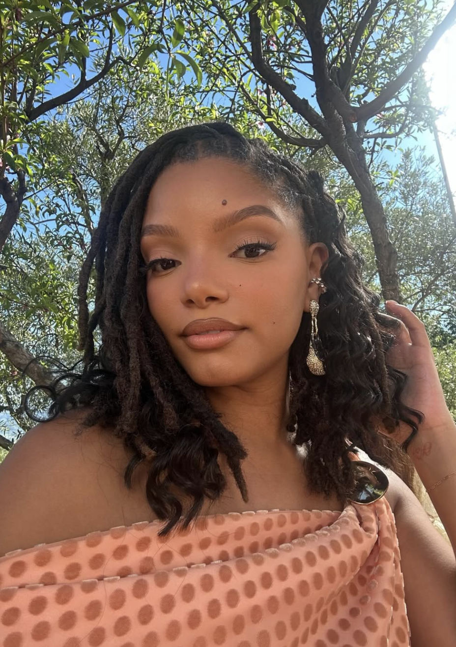 Halle Bailey with long, dark locs swept to the side, wearing an off-the-shoulder patterned top and showcasing how length and volume balance a heart face shape.