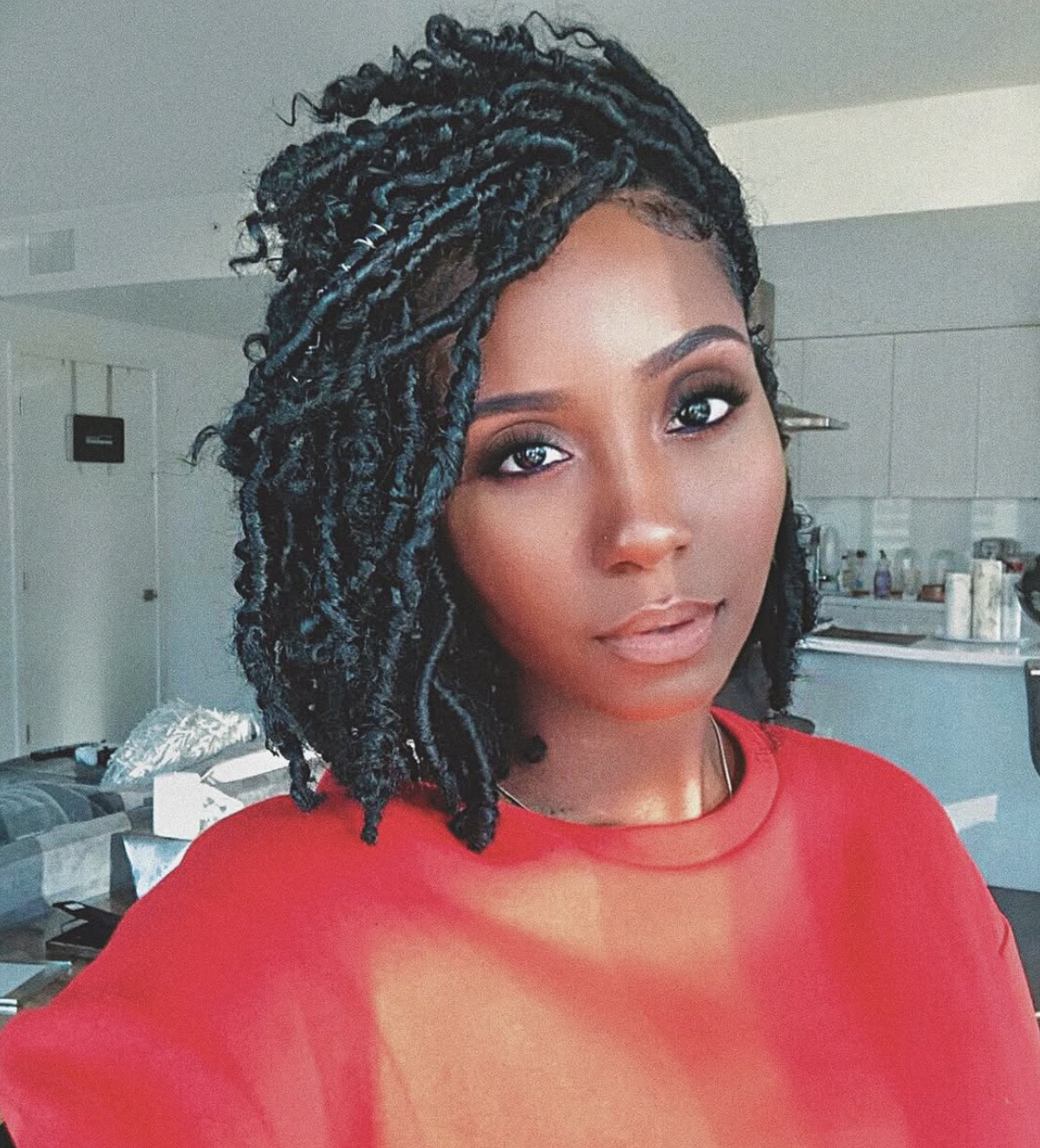 shoulder-length, textured faux locs and a side part, wearing a red top and showing how volume at the jawline balances a heart face shape.
