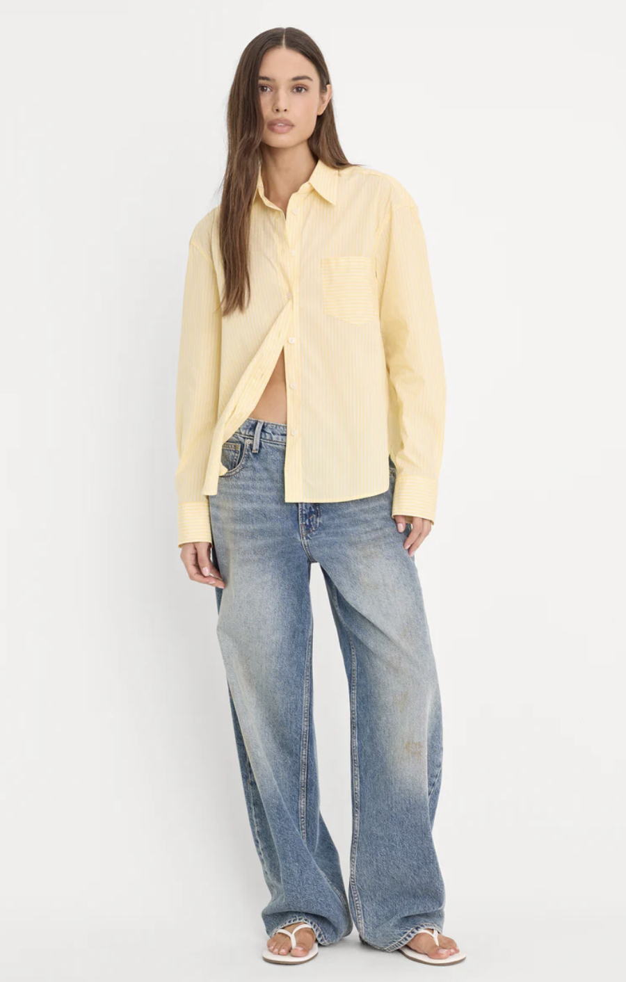 A close-up view of a person wearing an oversized light yellow button-down shirt unbuttoned at the waist, styled with loose wide-leg blue jeans and white flat thong sandals.