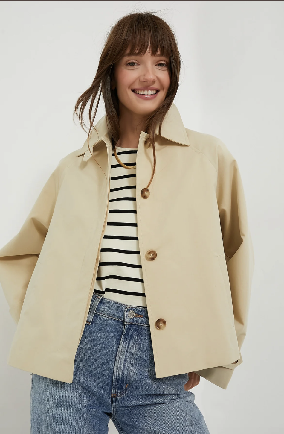 Cropped tan trench coat styled over a black and white striped top and high-waisted blue jeans.