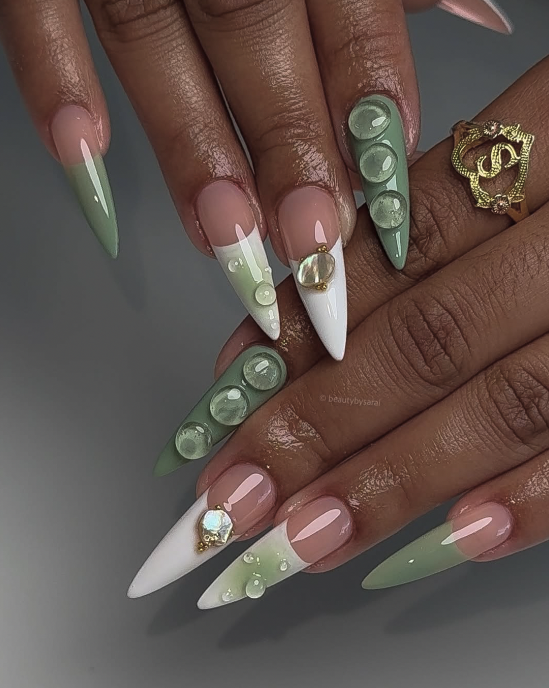 Long almond-shaped nails with a muted sage green base, hand-painted white flowers, sparkling crystals, and 3D clear gel water droplet accents.
