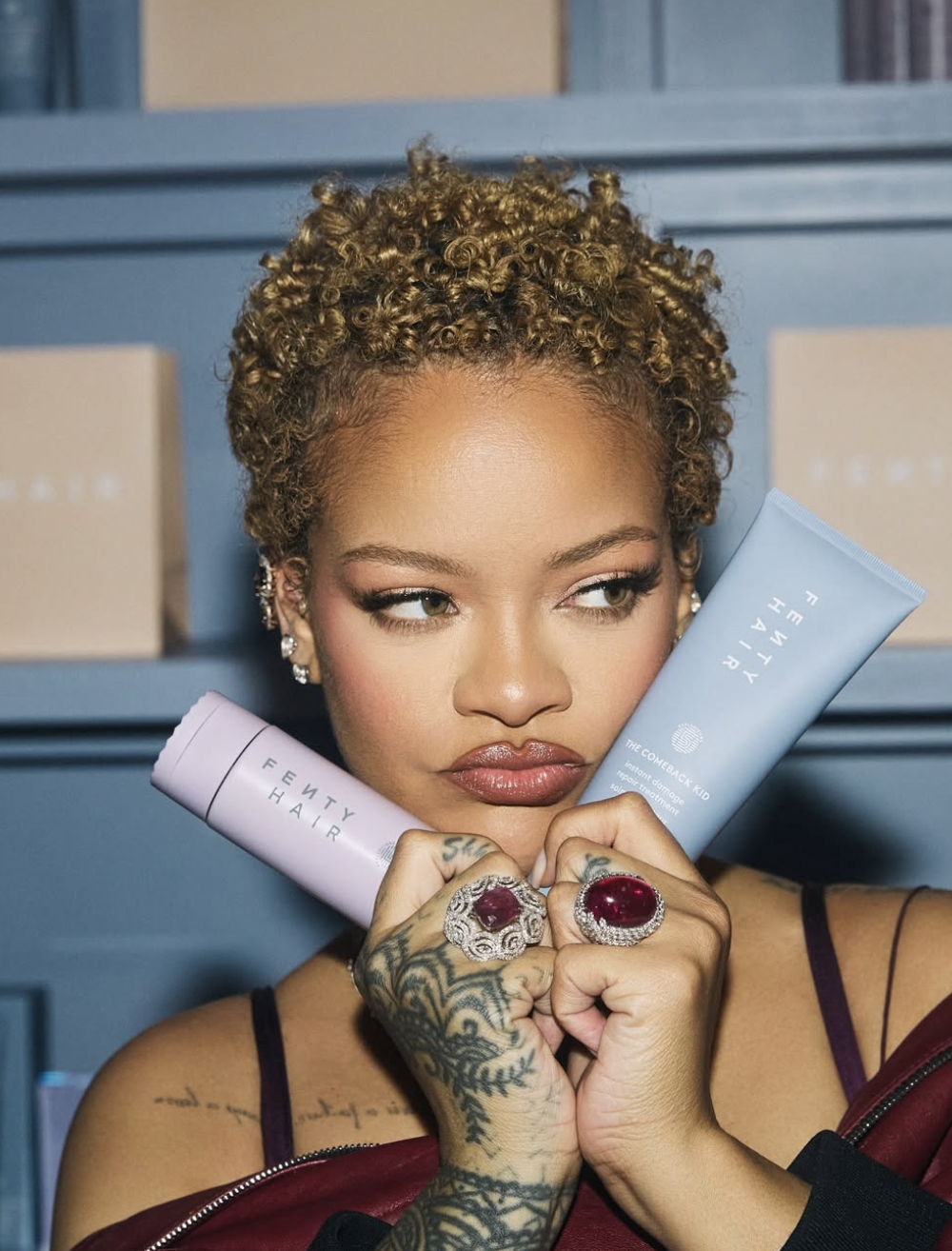 Rihanna showcasing a short, curly honey-blonde hairstyle while holding Fenty Hair products and wearing large ruby rings.