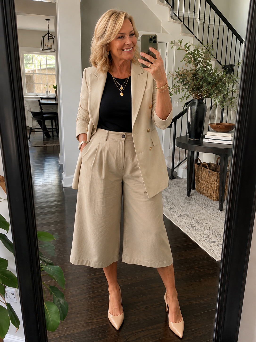 A woman taking a mirror selfie styling a monochromatic office-casual look with high-waisted, wide-leg beige linen shorts, a matching tailored blazer, a black top, and layered gold necklaces.