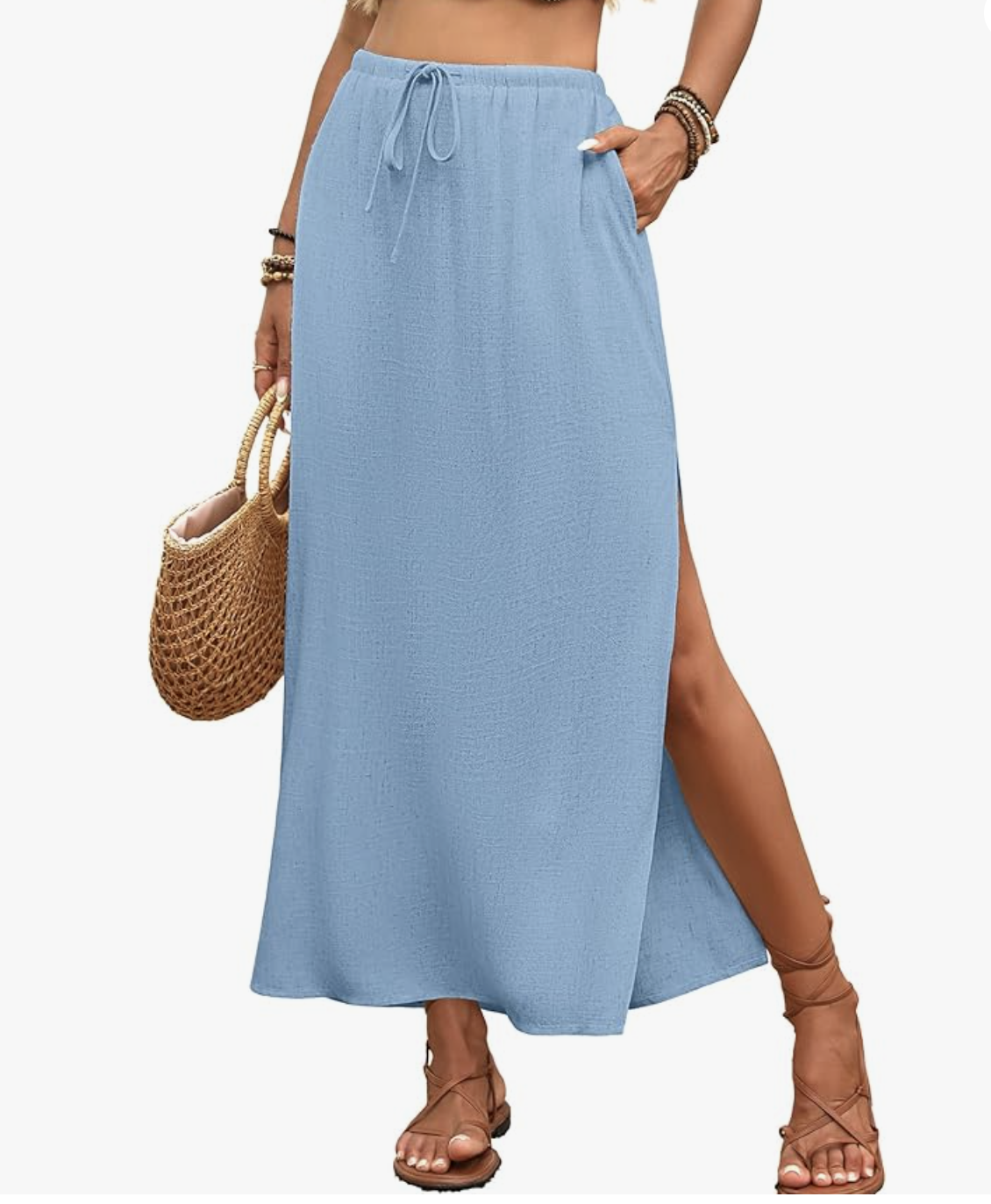 Close-up of a light blue linen straight maxi skirt with a side slit, styled with brown strappy flat sandals and a woven straw tote bag.
