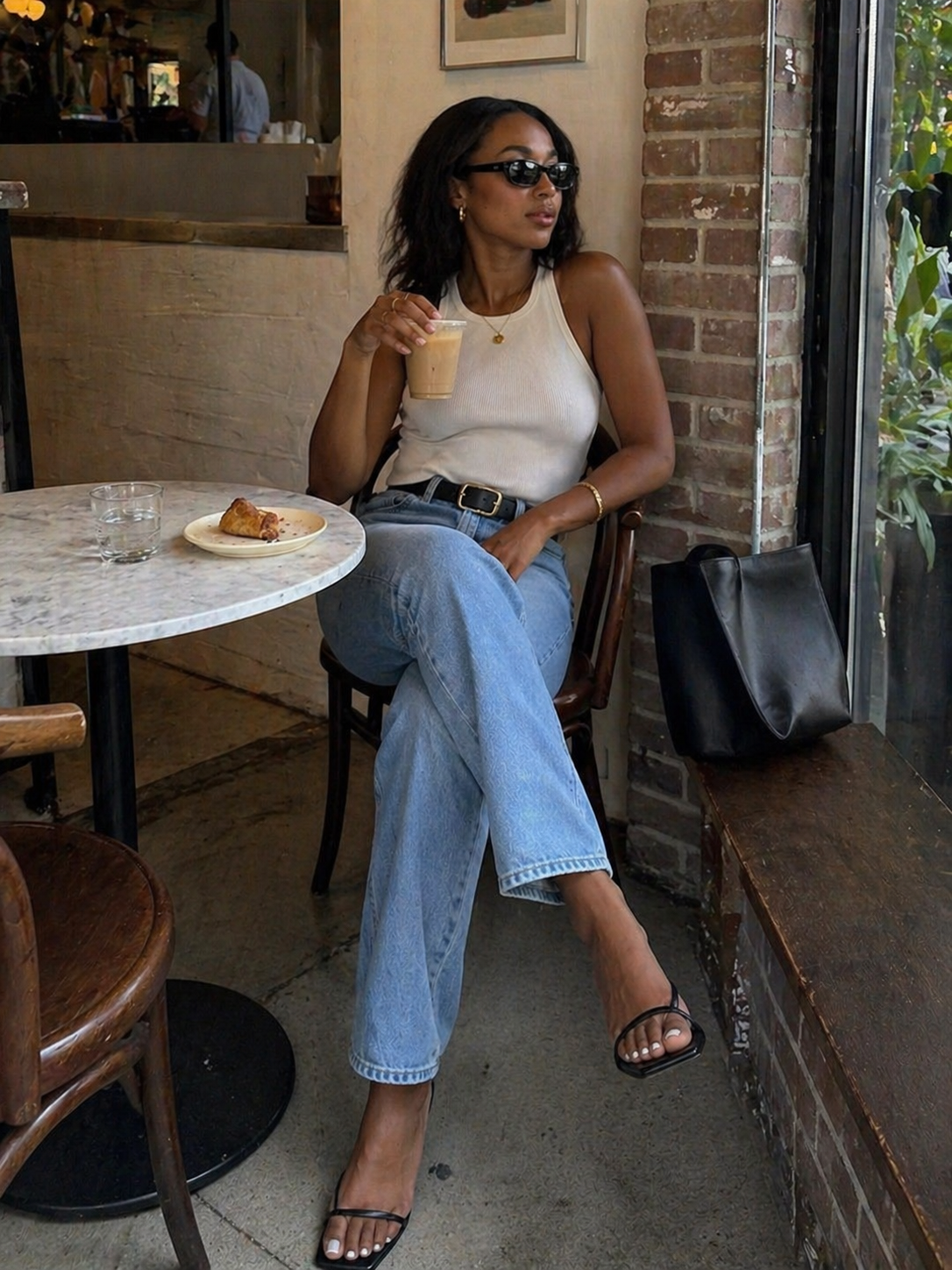 A woman sitting at a cafe wearing a white ribbed scoop-neck tank top tucked into light-wash straight-leg jeans with black strappy sandals.
