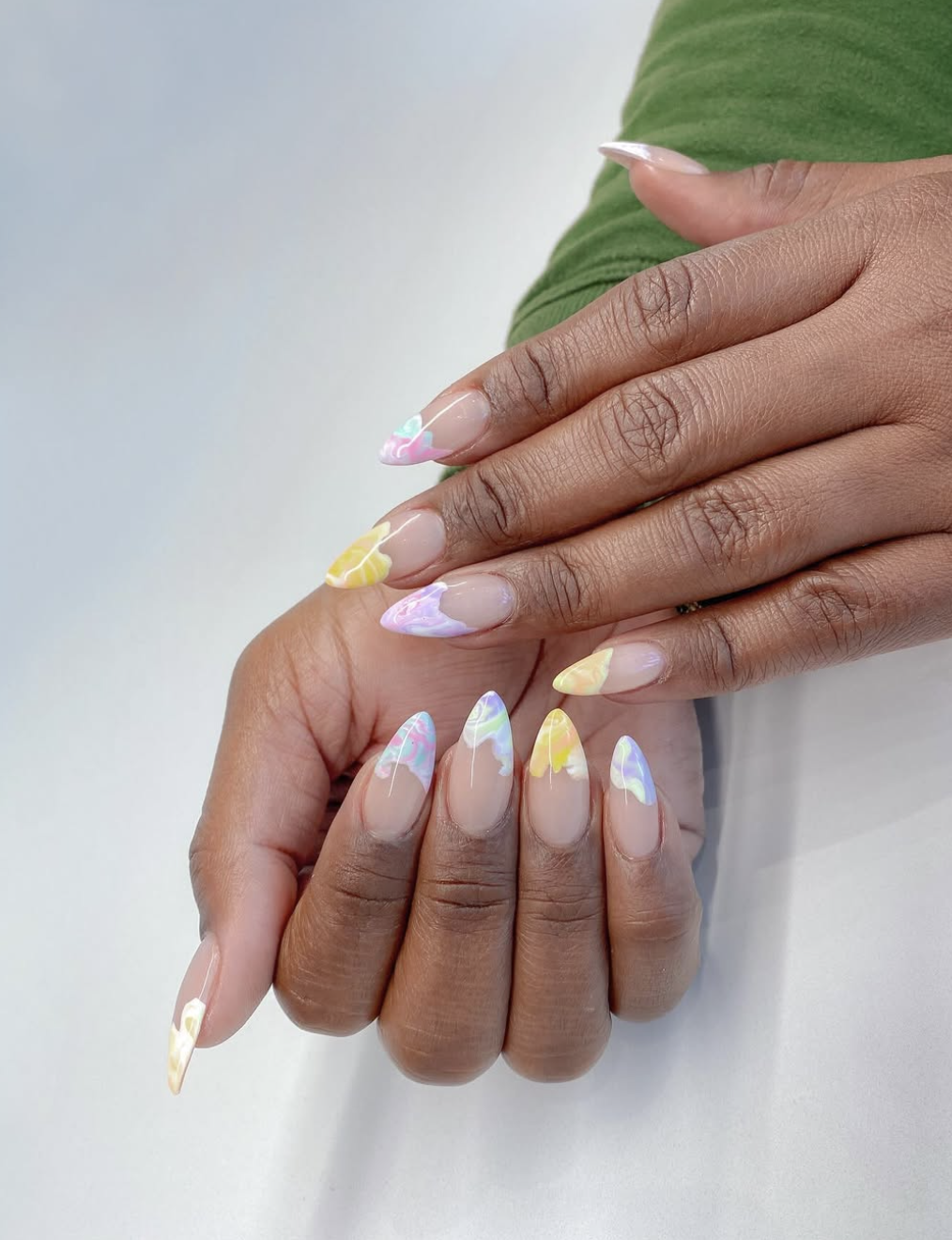 A close-up of a hand with long almond-shaped nails featuring a "pastel marble" design. The nails have a sheer base with airy, watercolor-inspired swirls of lavender, mint green, and butter yellow.