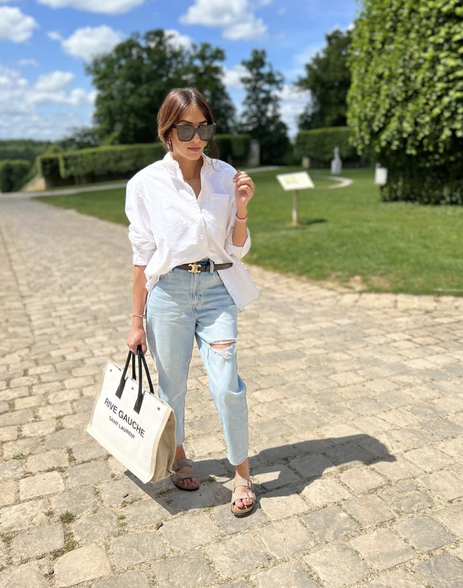 Woman walking on a cobblestone street wearing a white button-down shirt, light blue cigarette jeans, and slide sandals while carrying a canvas tote bag.