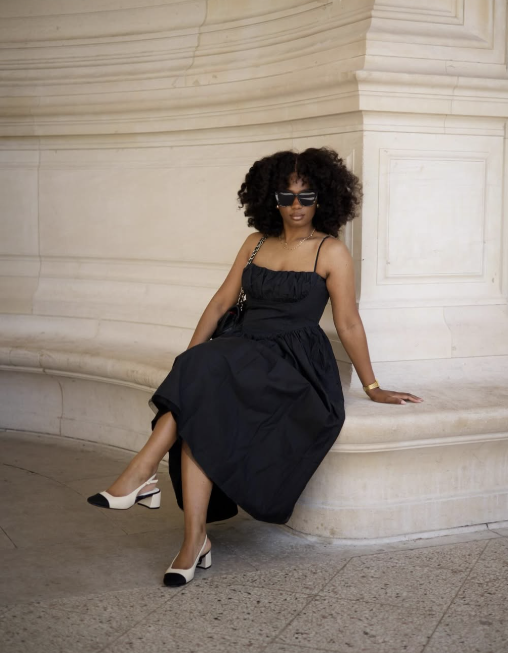 A woman with voluminous curly hair wearing a black strapless midi dress and two-tone cap-toe Mary Jane flats for women with a block heel, sitting against a stone pillar.
