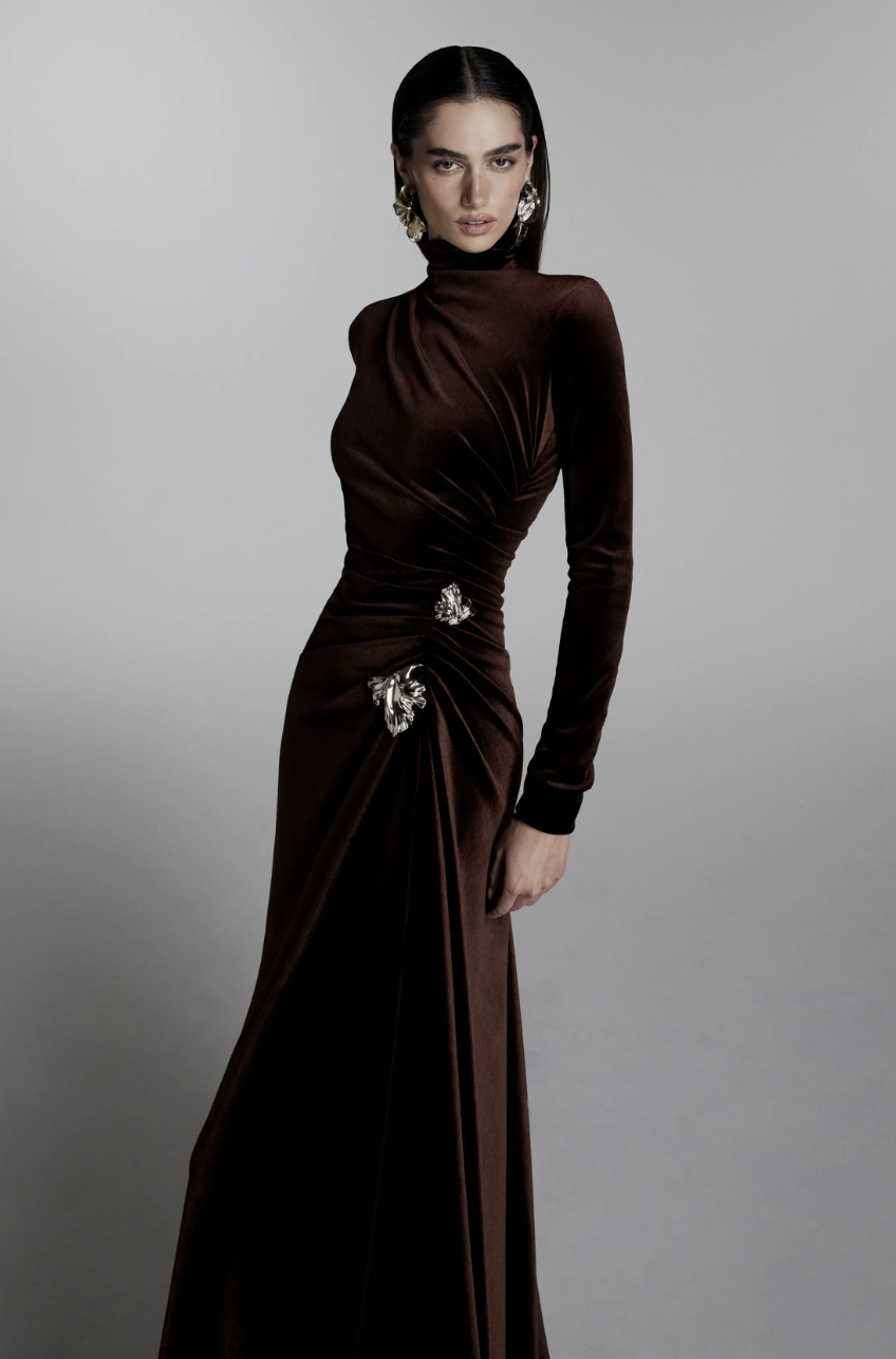 A model on the Monique Lhuillier Fall/Winter 2026 runway wearing a floor-length liquid velvet gown in deep chocolate brown, highlighting the season's warm luxury trend.