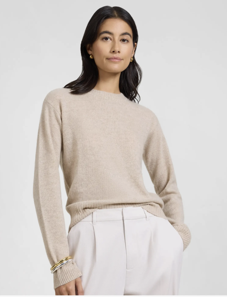 A woman wearing the Onia Featherweight Cashmere Crewneck Sweater in a neutral oatmeal color, tucked into white high-waisted trousers for a "Quiet Money" aesthetic.