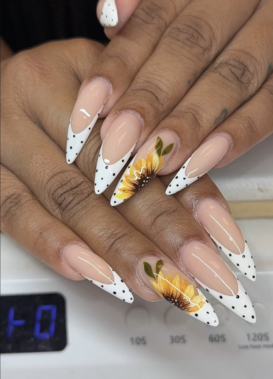 Stiletto-shaped nails with a white French tip and black polka dots, featuring a detailed yellow sunflower hand-painted on the ring finger.