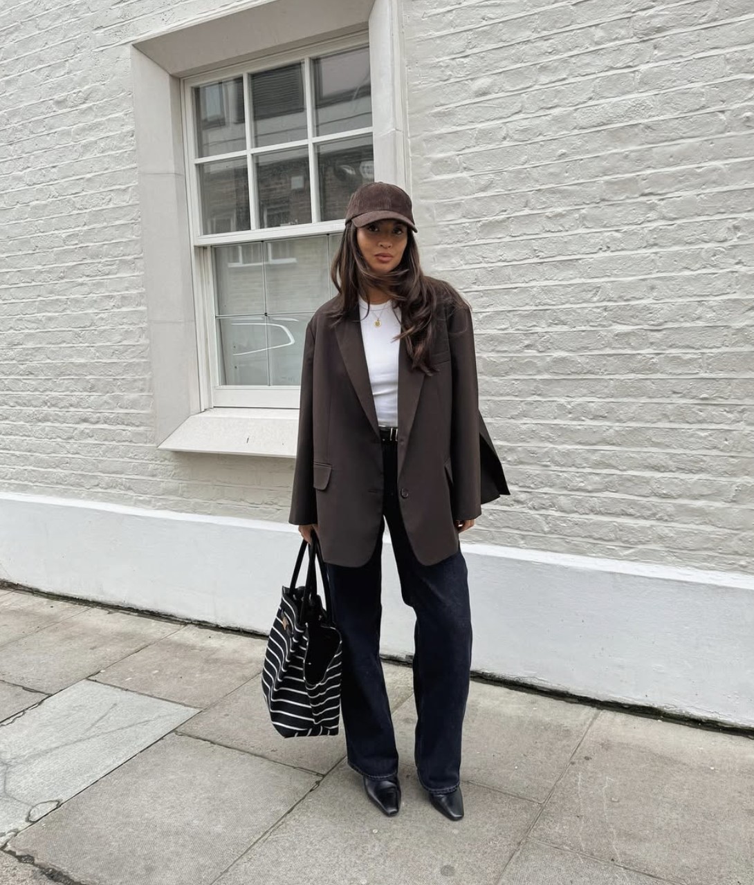 A stylist wearing an oversized chocolate brown blazer styled with dark-wash jeans, a striped tote bag, and a baseball cap for a polished everyday look.