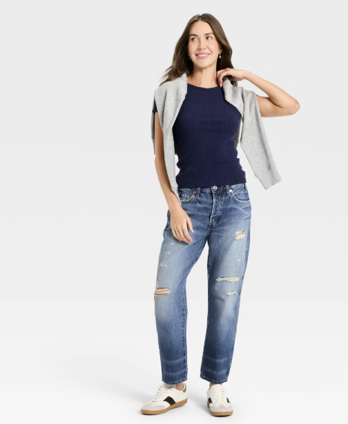 Model wearing Universal Thread Mid-Rise Boyfriend Jeans in a medium wash with rolled cuffs, styled with a navy blue mock neck top and a grey sweater over the shoulders to showcase a relaxed fit for an athletic figure.