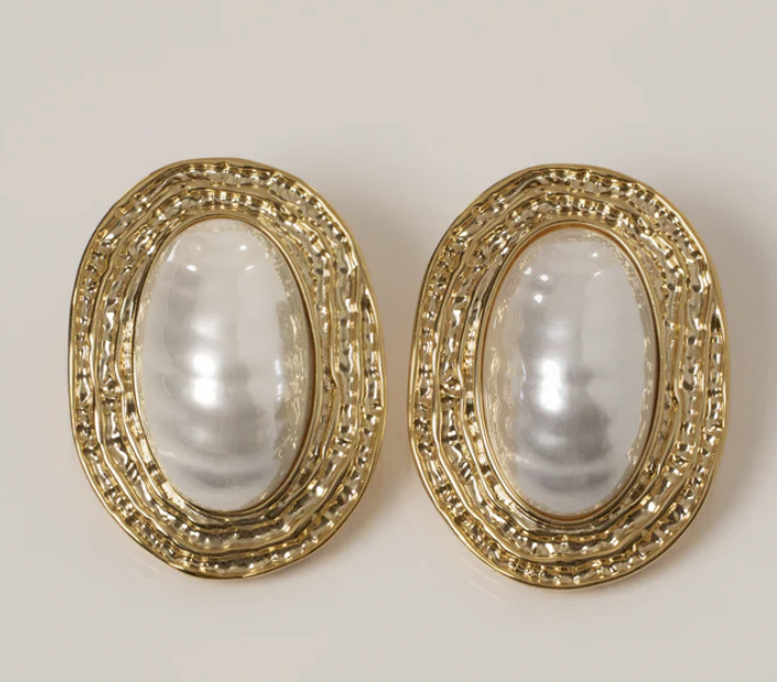 A pair of oval-shaped gold textured earrings with a large white pearl center, styled as a luxury accessory for a Mediterranean resort vacation.