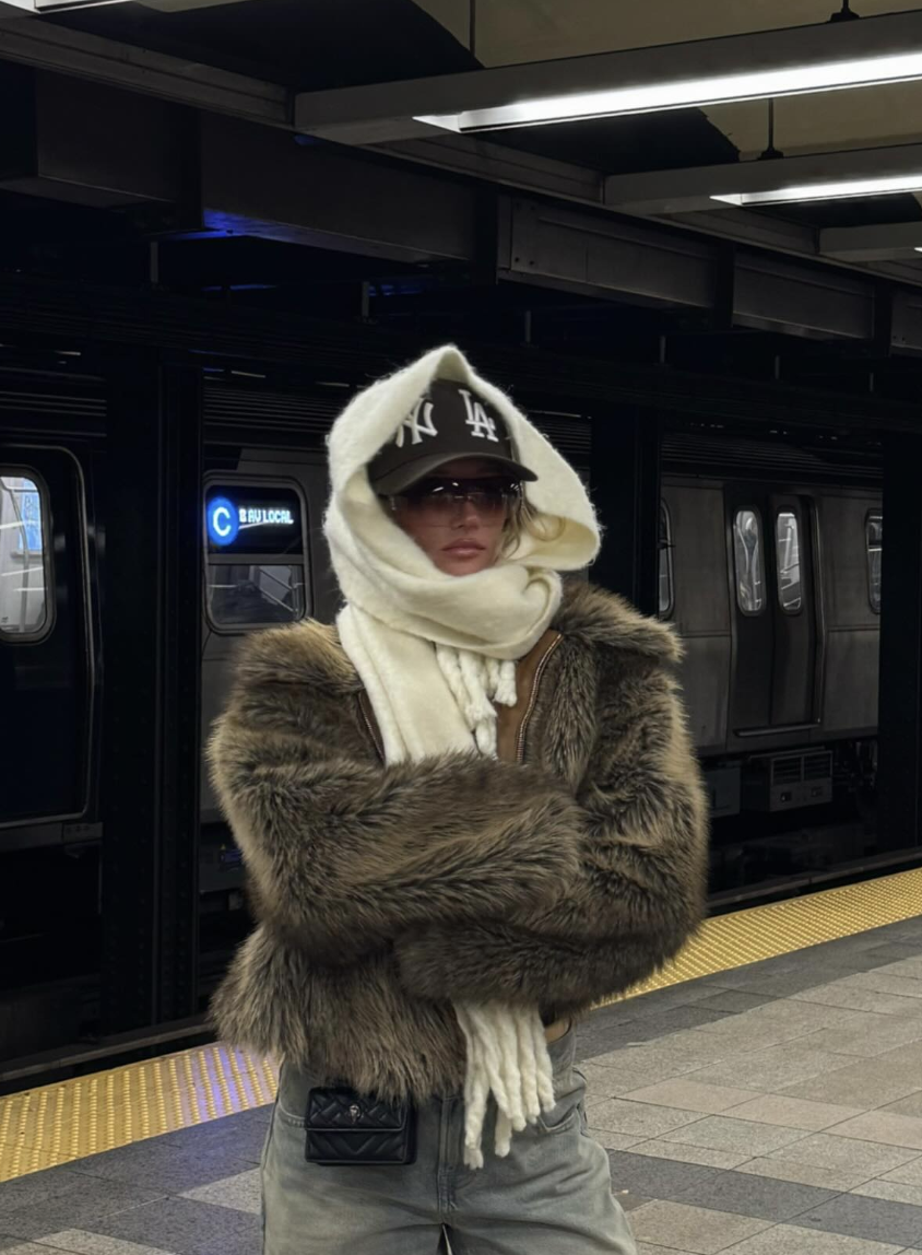 Brown furry-textured jacket with gray jeans, a cream scarf wrapped around the head and a dark baseball cap at a subway station.
