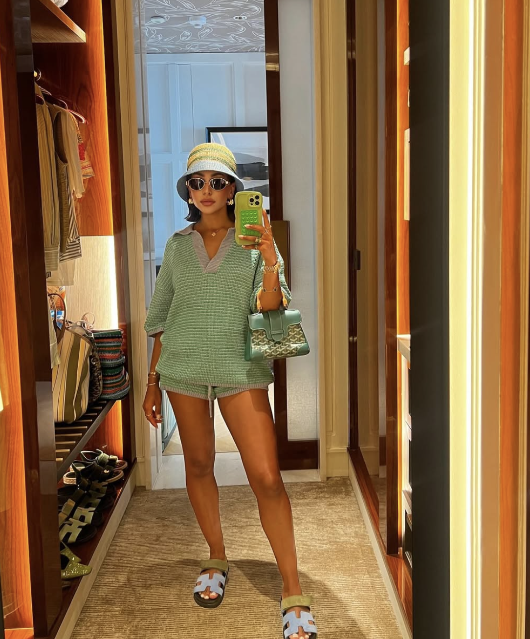 Mirror selfie of a woman wearing a casual, oversized light sage green knit polo shirt and matching short set, styled with a green patterned shoulder bag, oversized sunglasses, and a woven bucket hat for Summer 2026 style inspiration.
