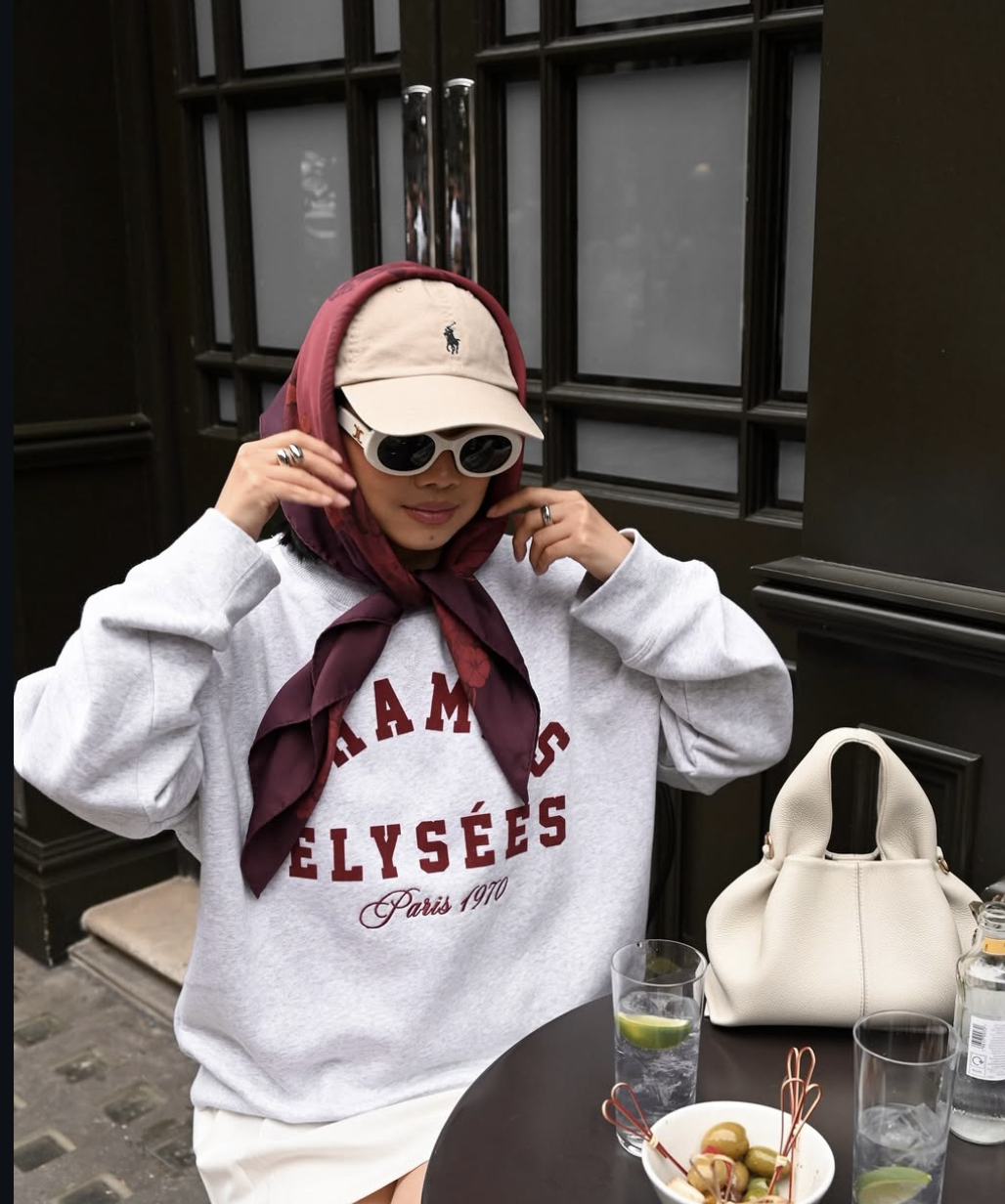 A close-up shot of a woman wearing a burgundy silk scarf wrapped over a beige Ralph Lauren baseball cap, paired with white oval sunglasses and an oversized grey 'Champs-Élysées Paris' sweatshirt.