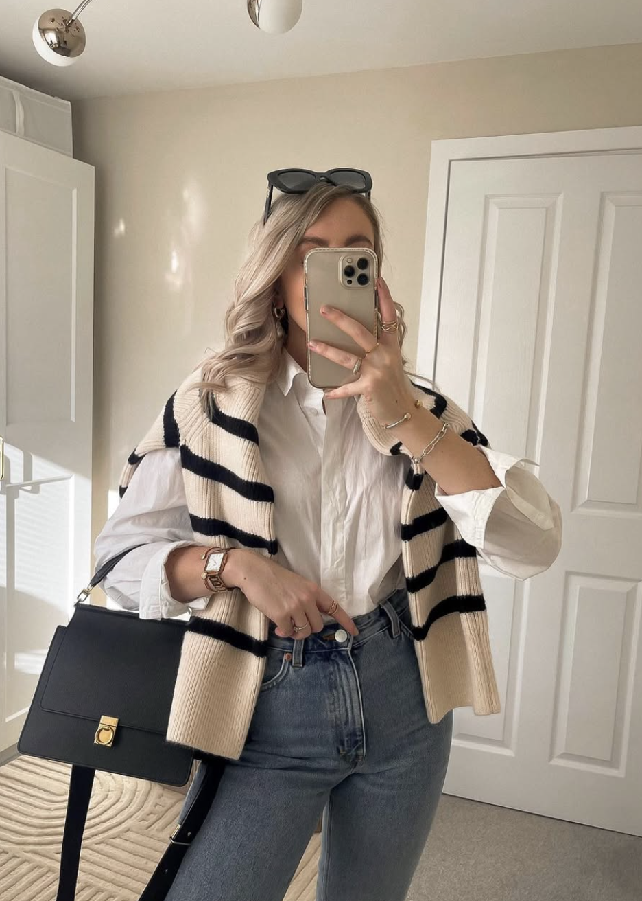 A woman wearing a white button-down shirt with a black and white striped sweater draped over her shoulders, paired with blue jeans and a black structured handbag.