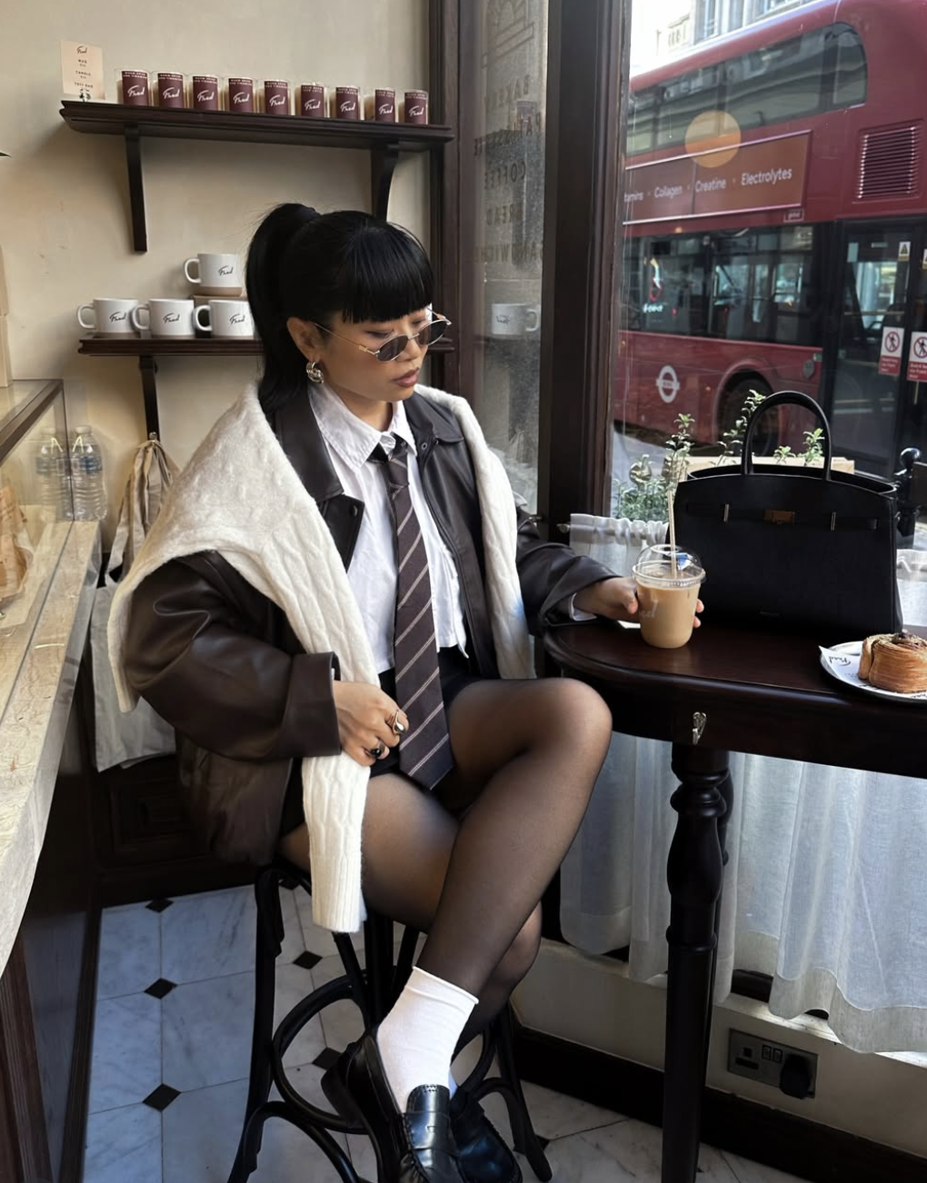 A woman wearing a dark brown leather jacket over a white button-down with a striped tie and sheer black tights, styled for a modern poetcore aesthetic.