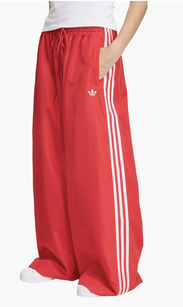 A close-up of a person wearing bright red adidas Originals Firebird Adilenium oversize track pants featuring the classic white three-stripe design and trefoil logo.